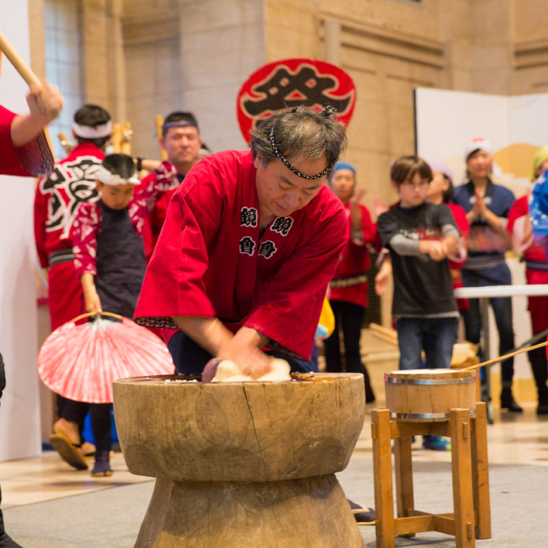 One person wearing a red top and a black headband kneads mochi dough while another person holding a mallet prepares to pound it.