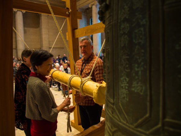 Three people hold wooden beam to ring a large bell